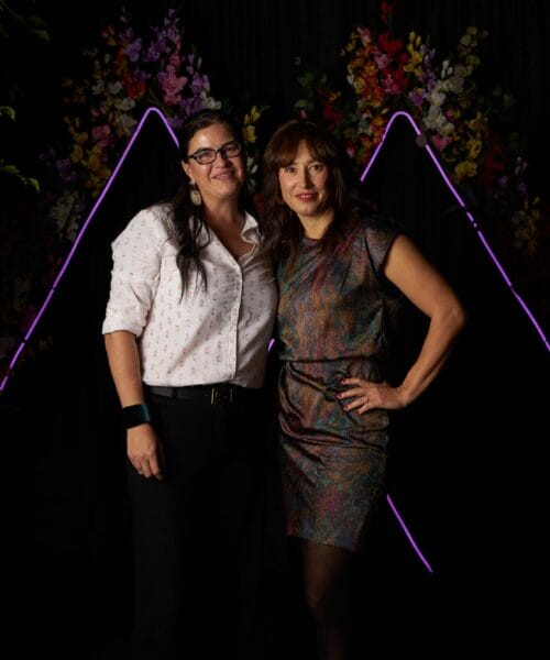 Two women pose in front of a dark backdrop decorated with colorful flowers and illuminated triangular neon lights. The woman on the left has long hair, glasses, and wears a white shirt with dark pants. The woman on the right has shoulder-length hair and wears a shiny metallic dress.
