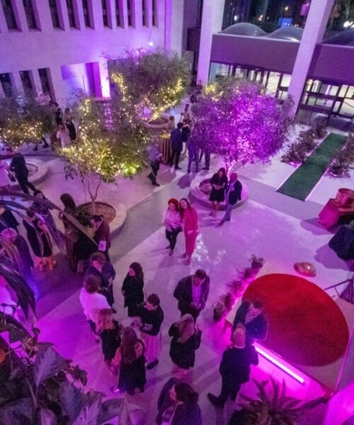 A spacious indoor venue with a high ceiling, decorated with lush greenery and purple lighting. Groups of people are gathered, some in conversation. A large screen is visible in the background, and there are various seating and table arrangements throughout the space.