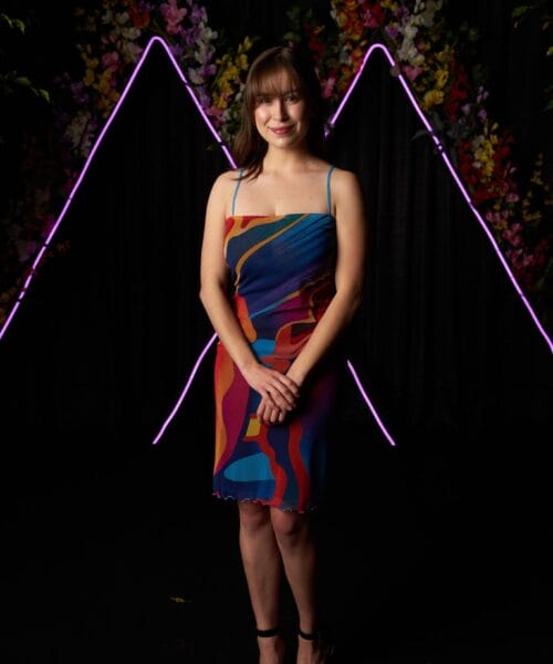 A woman stands in front of a vibrant backdrop featuring neon lights in the shape of inverted 