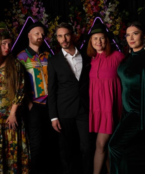 Five people posing together in front of a colorful, decorated backdrop. One person wears a floral dress with a leafy headpiece, another sports a vibrant geometric shirt and hat, a third is in a formal suit, and the remaining two wear a pink dress and a green outfit with a cape.