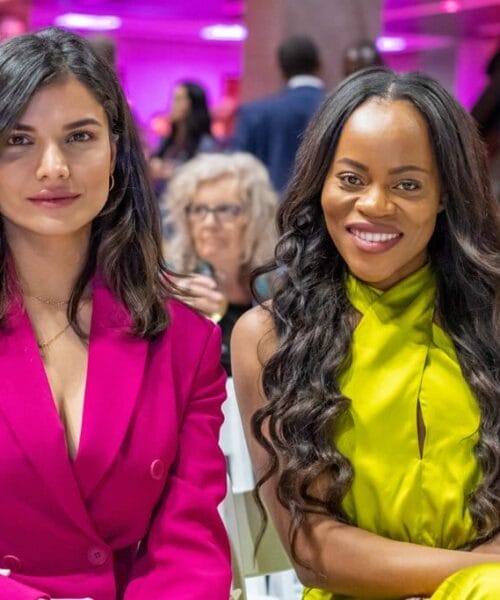 Two women sit together at an event. The woman on the left has shoulder-length dark hair and wears a bright pink blazer. The woman on the right has long curly hair and wears a bright green sleeveless top. They are surrounded by other event attendees in a brightly lit venue.