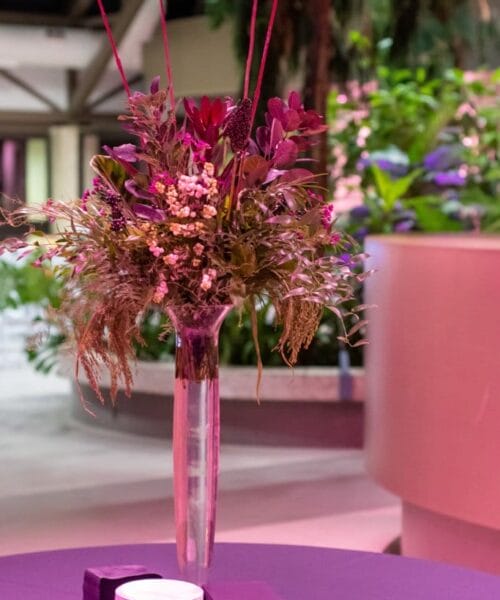 A tall, slender vase filled with an arrangement of purple flowers and greenery sits on a round, purple-covered table. Next to the vase are neatly stacked, small white plates and napkins. The background features lush, green plants and modern architectural elements, softly lit.