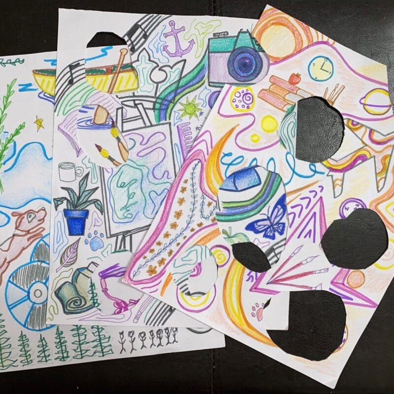 Three colorful pages filled with various doodles, including a rabbit, camera, rainbow, musical notes, and plants. Some areas are cut out, revealing holes in the paper. The background is a dark surface. The drawings are vibrant and whimsical.