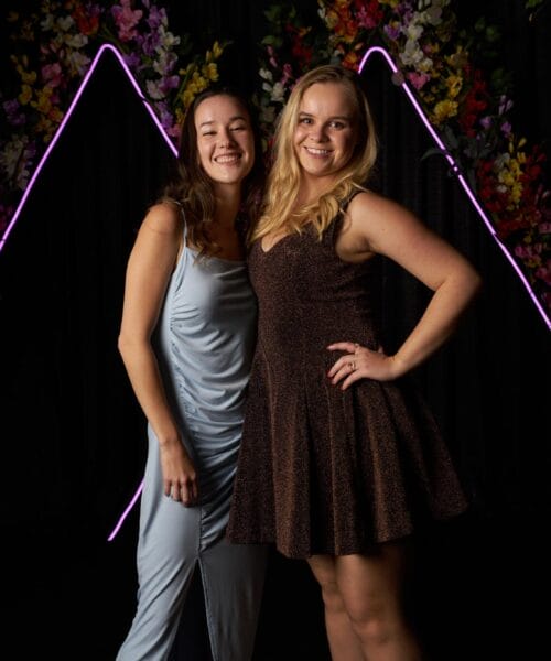 Two women are posing and smiling in front of a dark backdrop with purple neon triangle lights and decorative flowers. One woman is wearing a light blue dress and the other is wearing a brown sleeveless dress.