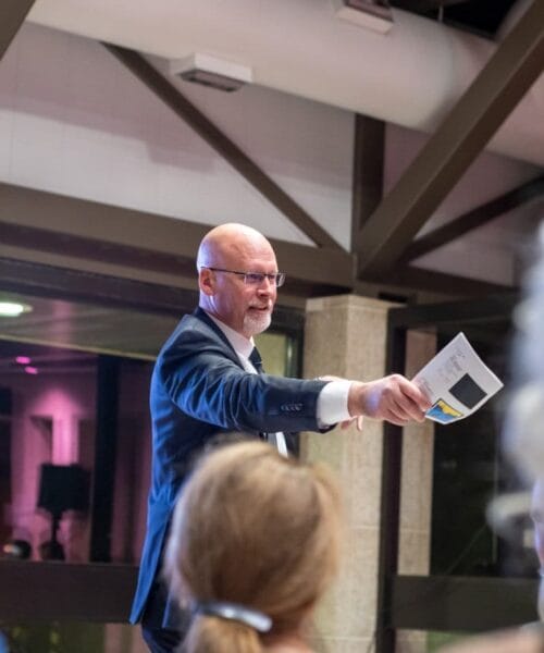 A man in a suit and tie is standing and gesturing with his right hand, holding a piece of paper. He appears to be speaking or presenting to an audience in an indoor setting with industrial-style beams and lighting. Several people are seated and facing him.