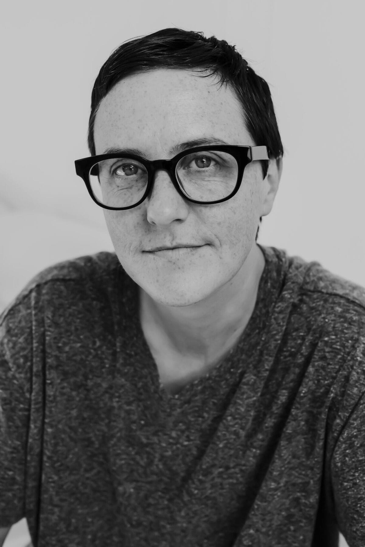 Gros plan en noir et blanc d'une personne aux cheveux courts, portant des lunettes à monture foncée et une chemise décontractée à motif texturé. Elle regarde directement la caméra avec une expression neutre. L'arrière-plan est légèrement flou.