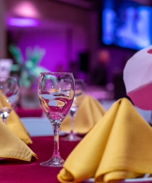A banquet table is elegantly set with folded yellow napkins, wine glasses, and a red tablecloth. The background is softly lit in purple, and large decorative pink flowers are visible. The ambiance suggests a formal event or celebration.