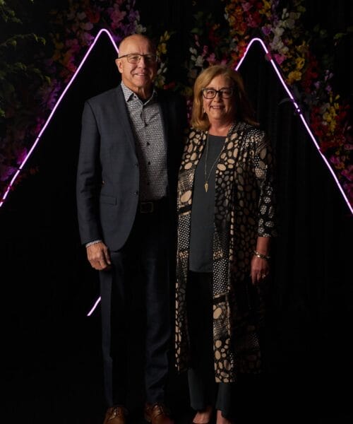 A man and a woman stand close together, smiling at the camera. The man is wearing a navy suit and patterned shirt, while the woman is wearing a patterned long coat over a dark outfit. They are in front of a dark backdrop with pink neon triangular shapes and colorful flowers.