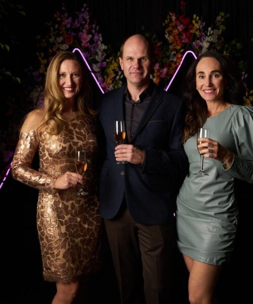 Three people dressed formally and holding glasses with drinks are posing for a photo in front of a backdrop with flowers and neon lights. Two women in elegant dresses are standing on either side of a man in a blazer and dress shirt.
