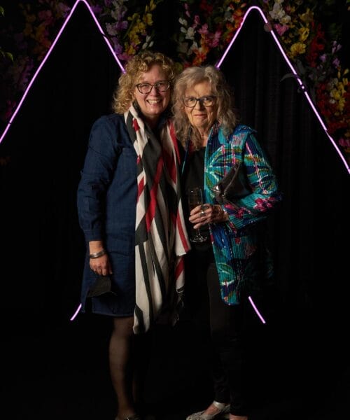 Two women stand close together, smiling at the camera. The woman on the left is wearing a dark dress, patterned scarf, and glasses. The woman on the right is in a colorful plaid jacket, holding a wine glass. They are in front of a decorative background with neon lights and flowers.