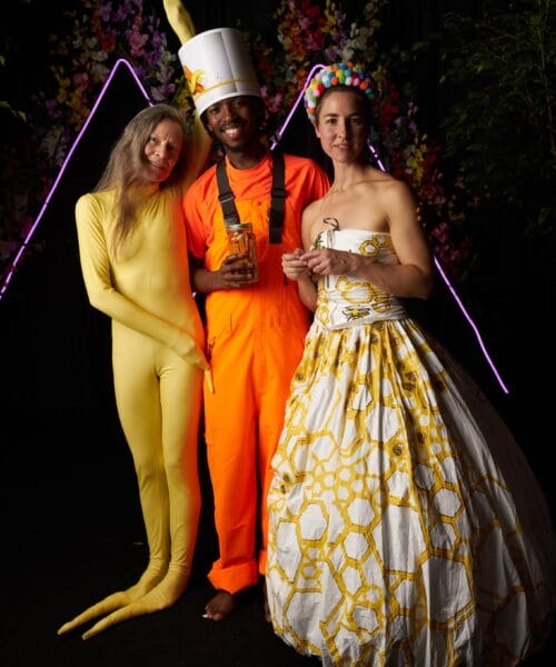 Three people are dressed in elaborate costumes. One person wears a yellow bodysuit and headpiece, another is in an orange jumpsuit holding a large head model, and the third wears a white dress with a yellow pattern and a colorful headband. They stand in front of a dark backdrop adorned with flowers and neon lights.