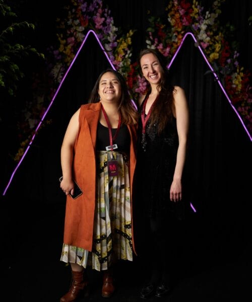 Two women are smiling and standing side by side in front of a backdrop adorned with colorful flowers and illuminated purple triangular light figures. One is wearing a long orange vest with a patterned dress, and the other is in a black dress. Both have event badges.