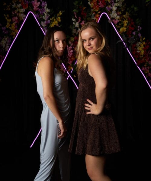 Two women pose confidently with their hands on their hips in front of a dark backdrop adorned with neon purple triangular outlines and vibrant flowers. The woman on the left wears a light blue dress, while the woman on the right wears a dark brown dress.