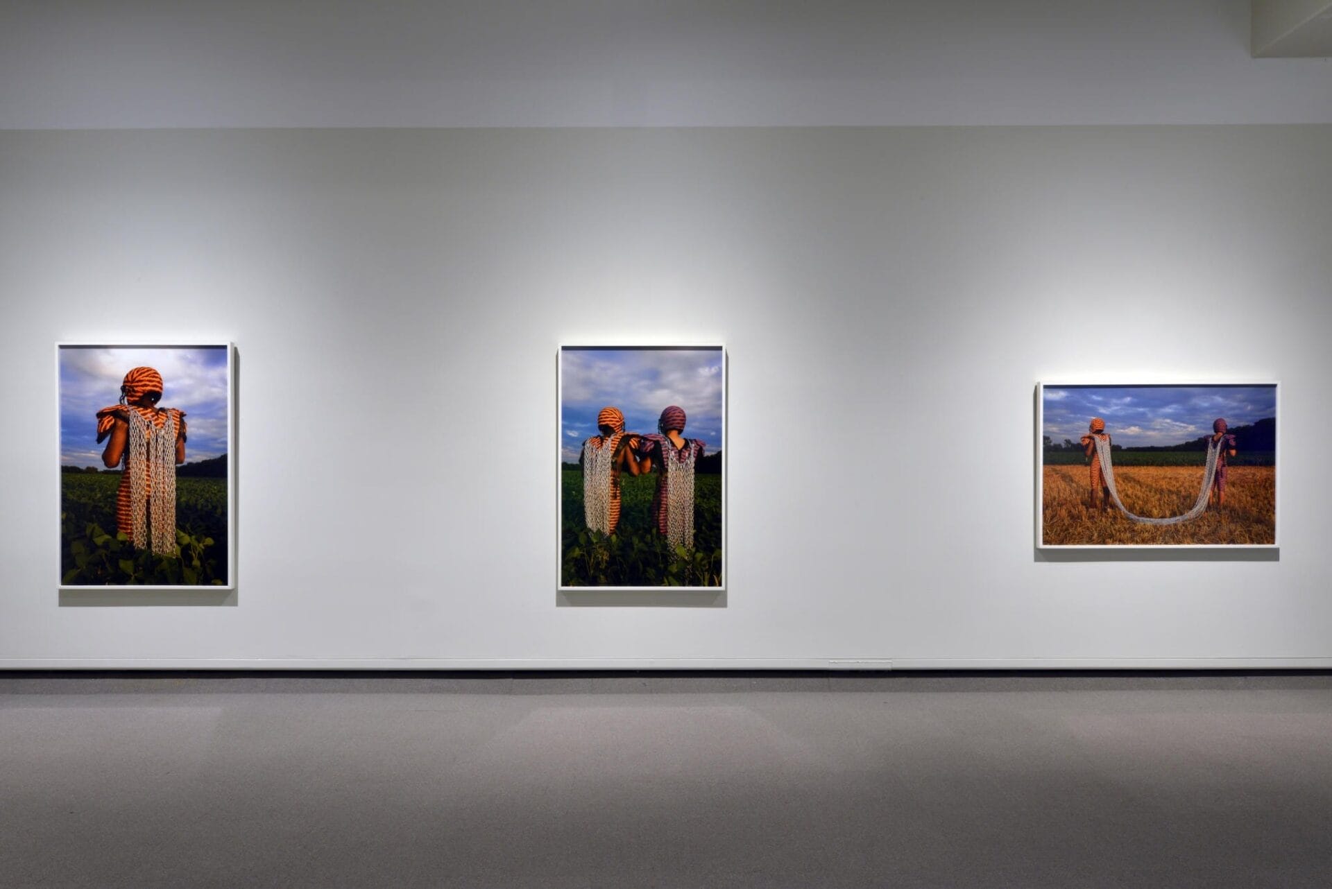 An art gallery wall with three framed photographs. The images depict figures in rural landscapes, adorned in colorful, woven textiles and headgear. The background features clear skies and fields. The photographs are evenly spaced and well-lit.