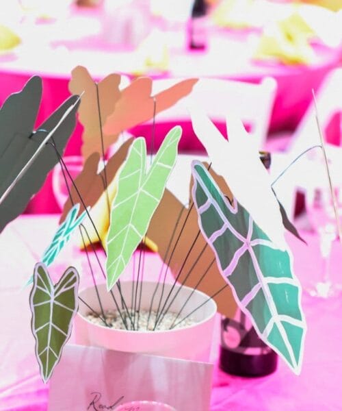 A white pot on a pink tablecloth holds various colorful leaf-shaped cutouts on sticks, serving as a vibrant table centerpiece. In the background, other tables with white chairs and decorations are visible, with a soft pink and white ambiance.