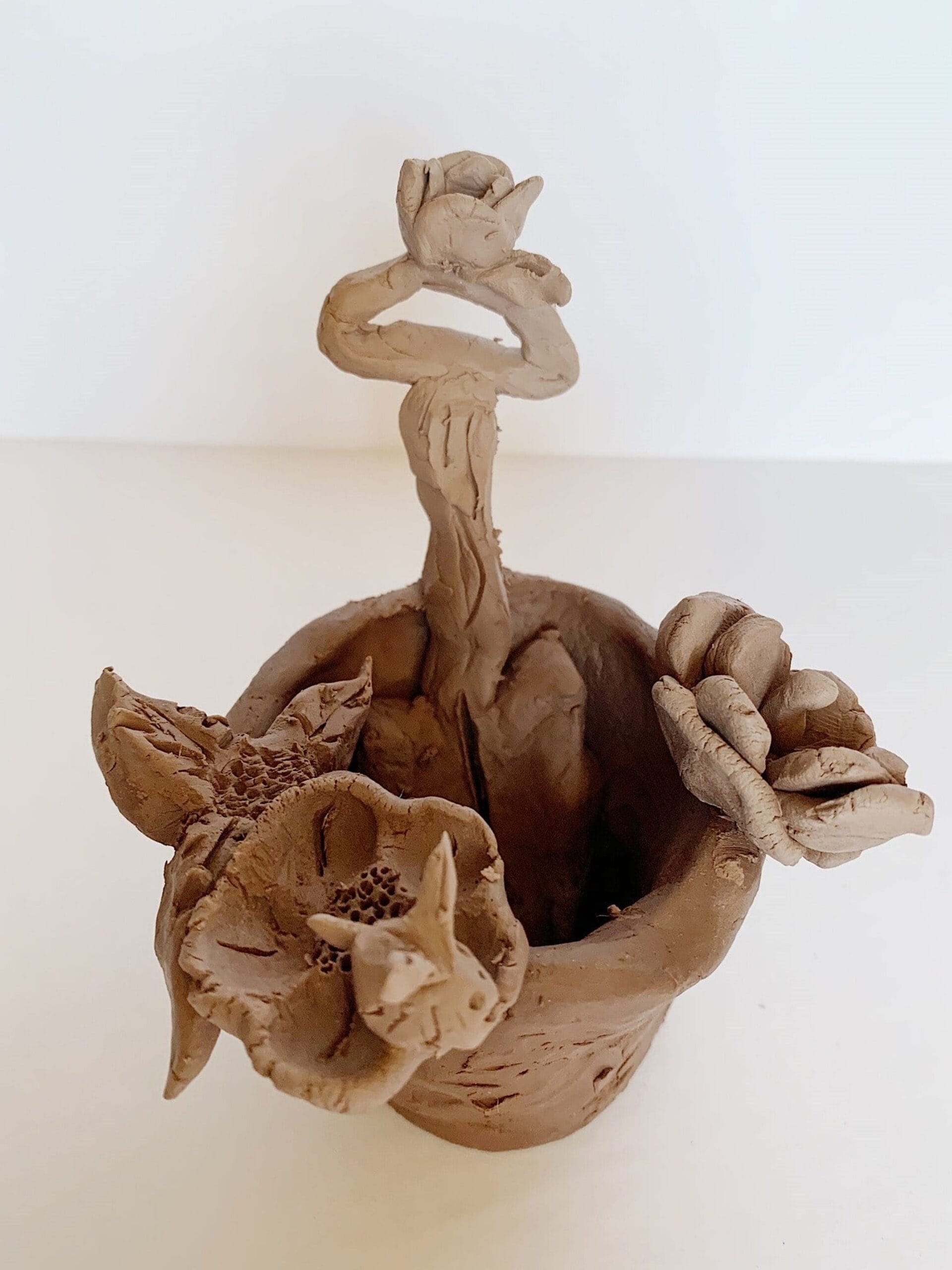 Une sculpture en argile fabriquée à la main représentant un petit pot avec des formes de fleurs détaillées qui en émergent. Les fleurs ont des textures variées et sont reliées par des tiges torsadées. La sculpture est placée sur un fond blanc uni.
