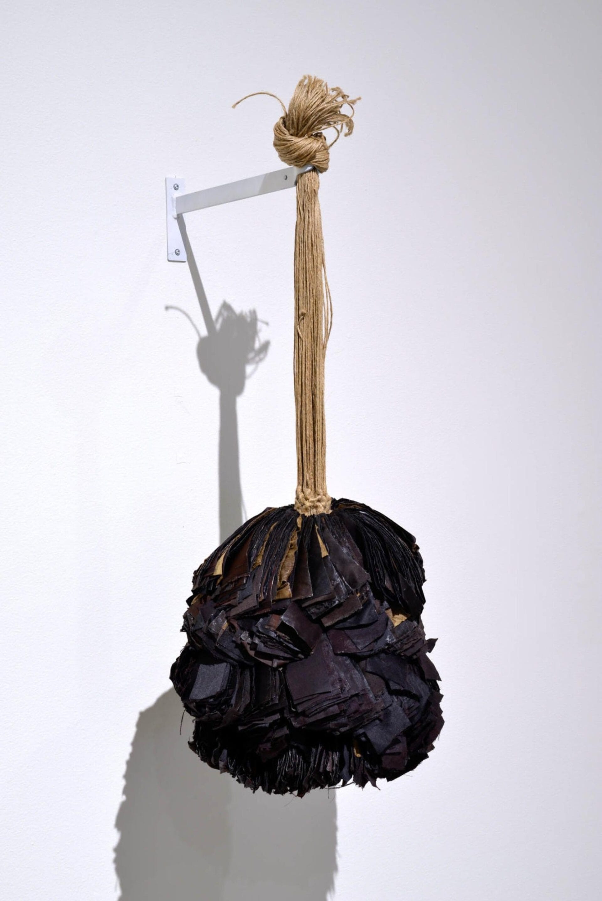 A contemporary art piece featuring a bundle of black textured material tied at the top with beige string. The bundle hangs from a white metal bracket extending from a wall, with its shadow cast on the wall behind it.