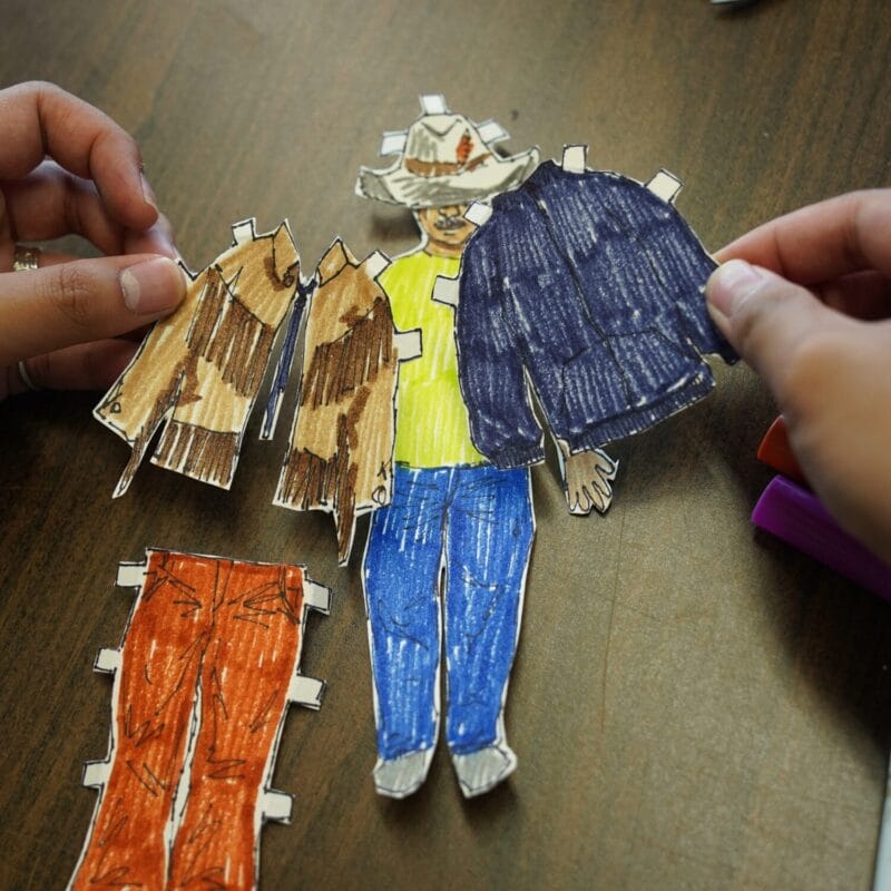 A person holds hand-drawn, colored paper cutouts of doll clothing, including jackets and pants, near a paper doll dressed in a yellow shirt, blue pants, and a cowboy hat on a brown table.