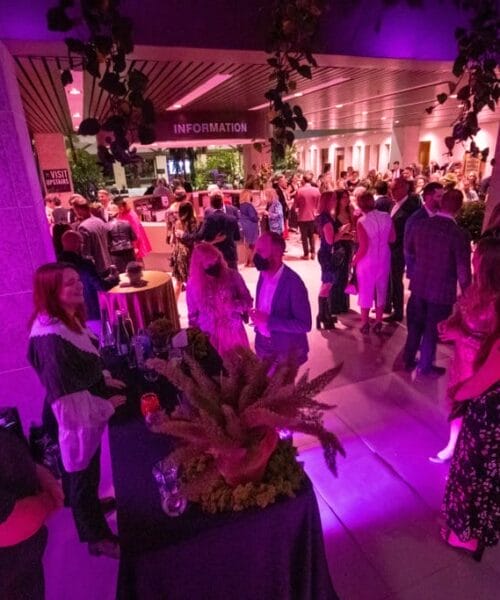 A large gathering of people is seen inside a building lit with vibrant pink lights. Guests are mingling, conversing, and enjoying drinks around tables adorned with decorative centerpieces. In the background, an 