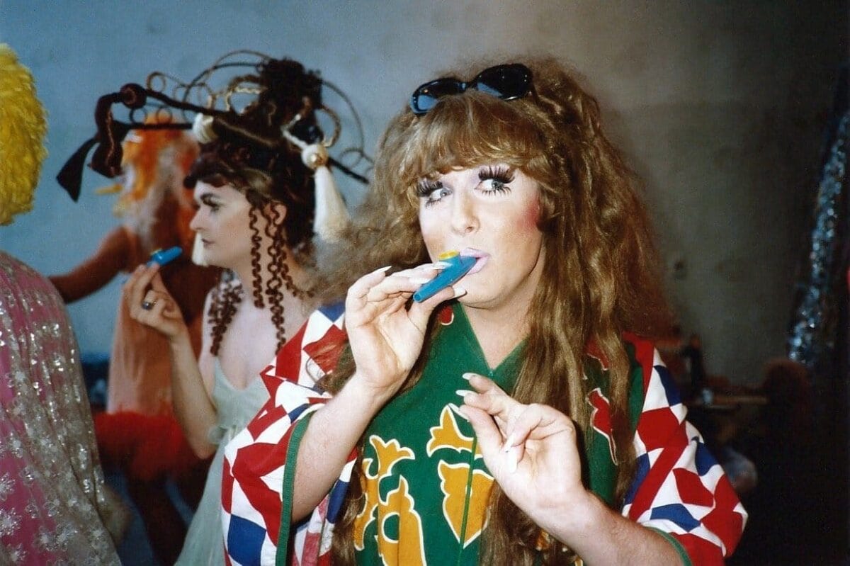A person with long curly hair, wearing sunglasses on their head and a colorful robe, holds a party blower to their mouth while others in creative costumes and elaborate headpieces are in the background. The setting appears to be an indoor gathering or event.