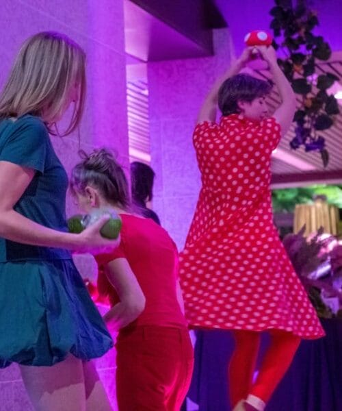 Three people are engaged in a lively performance under pink and purple lighting indoors. One person in a red polka dot dress is jumping with arms raised, holding a red and white prop. Behind them, someone in blue holds green objects. The background features other onlookers.