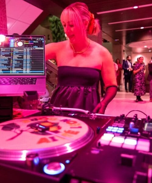A DJ stands behind a turntable and mixer, with a laptop displaying a music playlist. The DJ is wearing a strapless dress and gloves. Guests are seen mingling in the background at an indoor event with pink and purple lighting.
