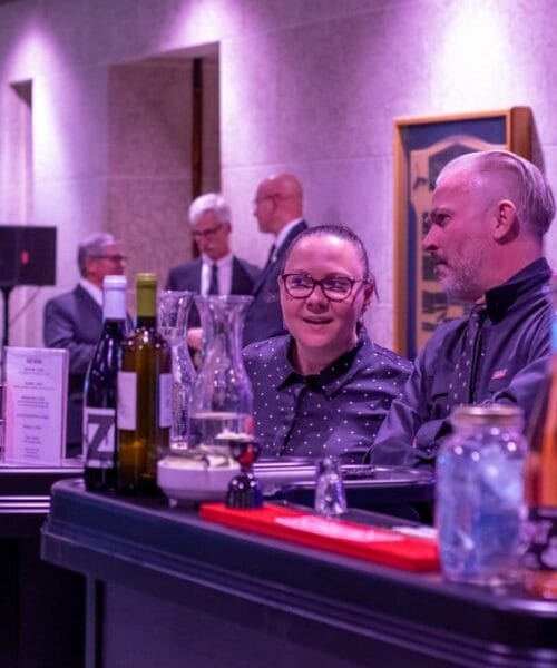 Two people chat at a bar counter in a purple-lit room. The counter is lined with various bottles, glasses, and cocktail shakers. In the background, several individuals are conversing near a wall adorned with artwork.