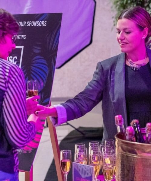 At a reception or event, a woman holding a bottle of sparkling wine pours a glass for another individual. Around them, a table is set with multiple glasses of champagne and a bucket holding additional bottles. The lighting casts a pinkish hue over the scene.