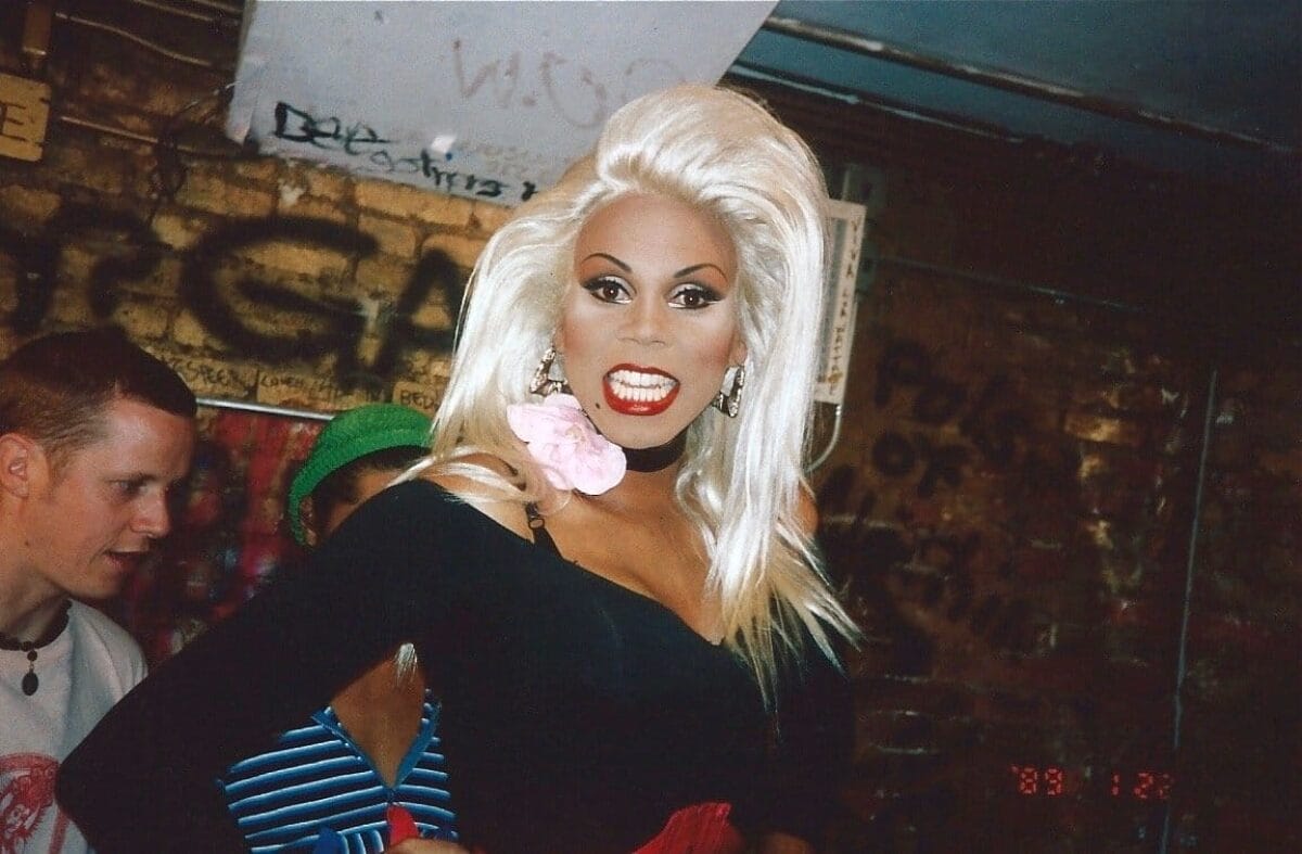 A person with long blond hair, dramatic makeup, and a black outfit with a pink flower poses confidently in front of a graffiti-covered wall. Two other individuals are partially visible in the background, one wearing a hat and another wearing a striped shirt.