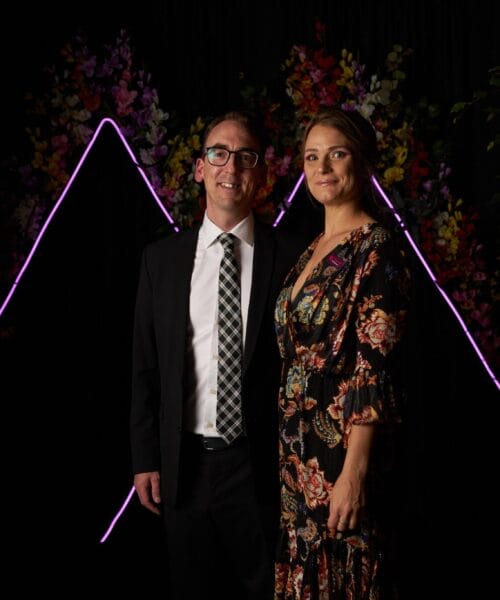 A man in a suit with a checkered tie and glasses stands next to a woman in a floral dress. They pose in front of a dark backdrop adorned with purple neon lights forming a 'W' shape and various floral arrangements. Shrubs frame both sides of the scene.