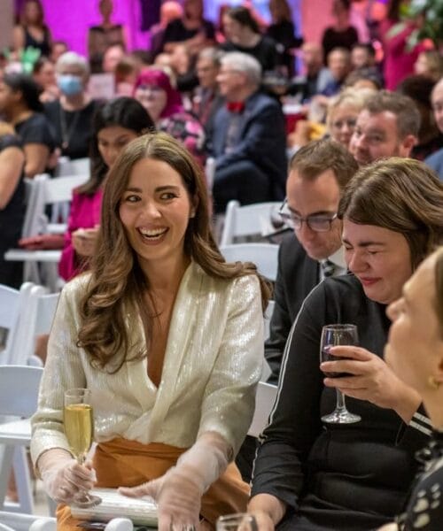 A woman with long brown hair and a white blouse is smiling and holding a glass of champagne while sitting among a crowd at an indoor event. People around her are also engaged, holding drinks and conversing, as they sit on white chairs. The background is brightly lit.