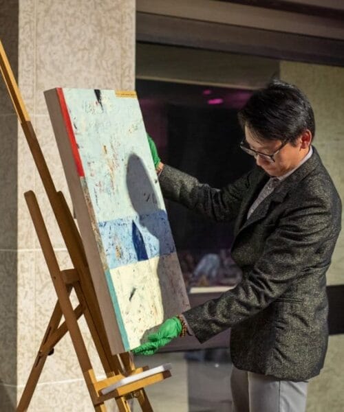 A person wearing a dark suit and green gloves is holding a painting on an easel, examining it closely. Another person in a suit stands beside a podium, speaking into a microphone. They are indoors, in a room with glass windows and walls.