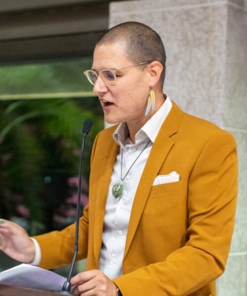 A person with short hair, glasses, and a yellow blazer speaks at a podium. They wear a white shirt, green pendant necklace, and white pocket square. One earring is visible. A microphone and some notes are present on the podium. Indoor setting.
