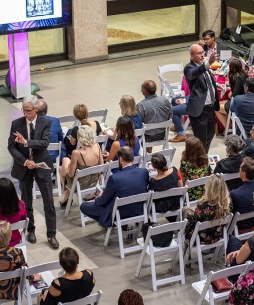 A man in a suit stands and gestures in front of a seated audience at an indoor event, possibly a seminar or conference. Attendees sit on white chairs, facing forward, with a screen displaying content in the background. The atmosphere appears formal and engaging.