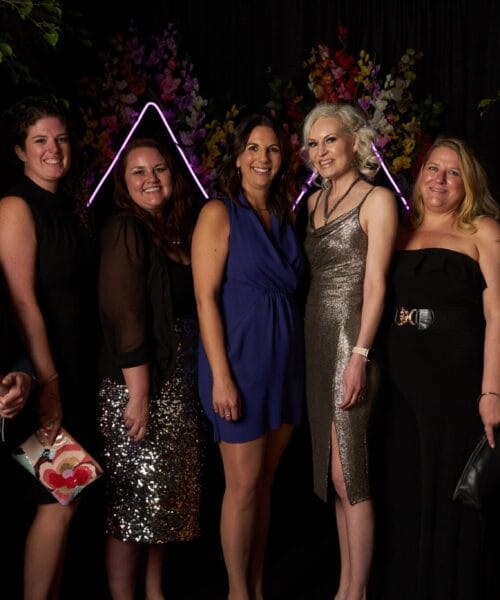 A group of seven people, four women and three men, pose for a photo. The women are wearing dresses in various colors, and the men are wearing shirts and trousers. They stand in front of a backdrop with illuminated triangular designs and floral decorations.