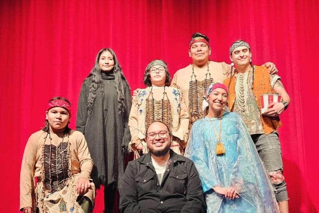 A group of seven people, some in traditional Indigenous attire, pose on a stage in front of red curtains during RAWLCO Free Day. Six stand or kneel, while one sits cross-legged in front, smiling at the camera.