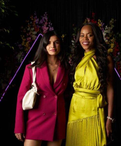 Two women stand side by side, smiling at the camera. The woman on the left wears a vibrant pink blazer and holds a white purse, while the woman on the right dons a yellow dress with pleated details. They're in front of a black backdrop adorned with floral decorations and purple neon accents.