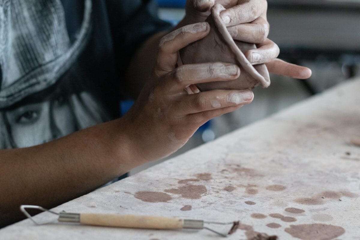 A person with clay-covered hands shapes a piece of clay on a worktable. Nearby, a clay sculpting tool rests on the table, which is also covered with clay spots. Focused on their work, they appear to be creating or molding a small pinch pot.