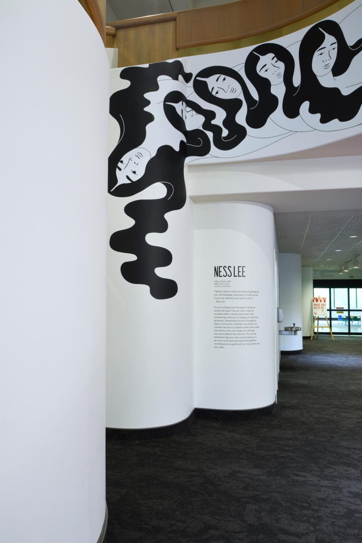 Wall art featuring stylized black and white illustrations of faces with flowing hair is displayed on a curved white surface. The adjoining wall includes a text description and the name 