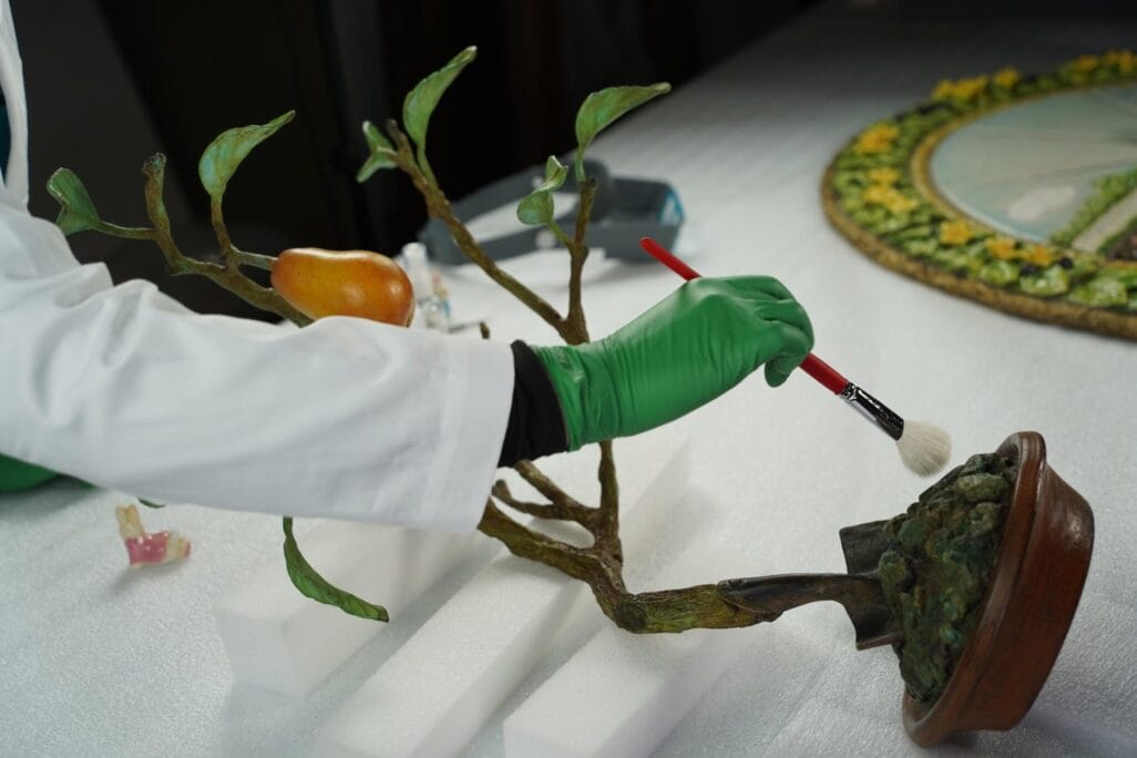 A person wearing green gloves and a white coat is using a brush to clean a small tree sculpture with a yellow fruit. The sculpture sits on a foam pad, and a decorative circular item is visible in the background.