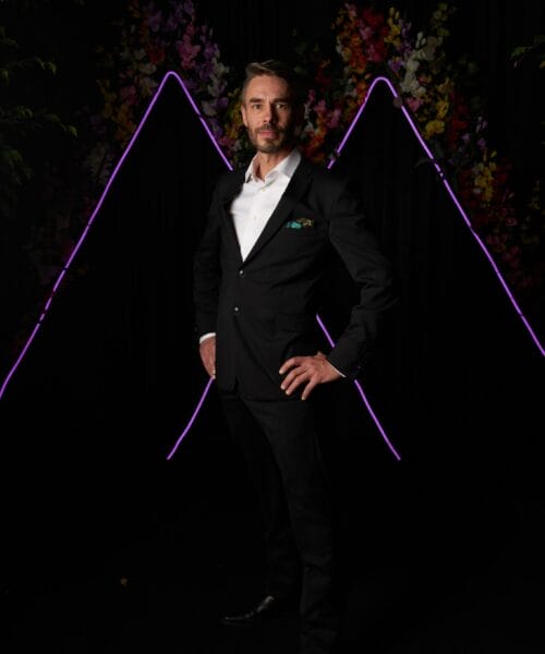A man with short hair and a beard stands confidently with one hand on his hip, wearing a black suit and white dress shirt. The background features a dark setting with vibrant, colorful foliage and two V-shaped pink neon lights.