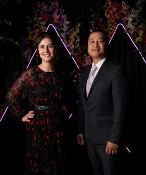 A woman in a black dress with red accents and a man in a dark suit stand side by side, smiling, in front of a backdrop with purple neon triangular lights and colorful flowers. Green foliage surrounds the backdrop.