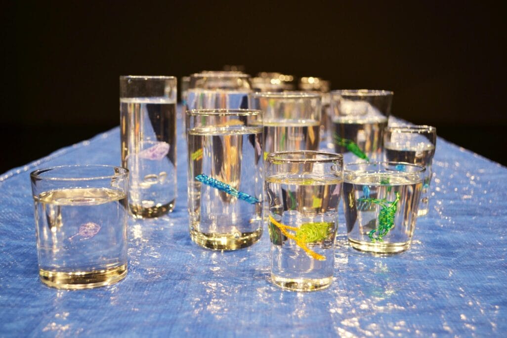 A collection of glass cups filled with water is placed on a blue surface. Each glass contains a small, colorful object resembling a sea creature, featuring various colors like green, blue, and yellow beads in the blood of the water.