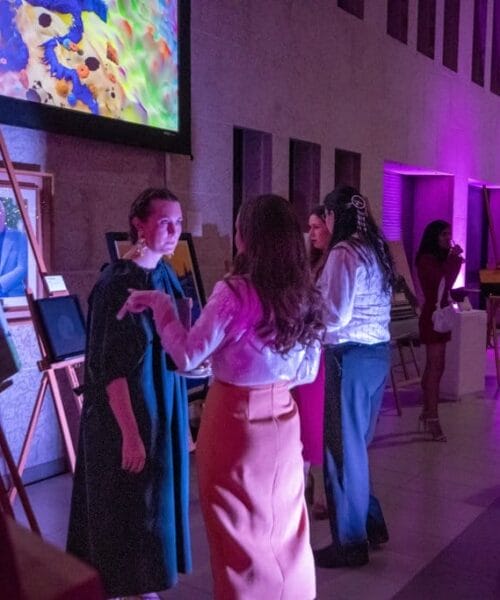 Several people gather indoors at an art exhibition, which is dimly lit with purple lighting. They are observing and discussing framed artworks displayed on easels. The walls and floor are light-colored, complementing the vibrant atmosphere.