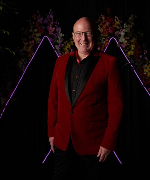 A man wearing a red suit jacket and black shirt is smiling while standing in front of a dark backdrop with neon purple triangular outlines and colorful flowers. Green foliage is partially visible on the left and right sides.