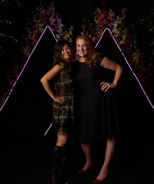 Two women are standing and smiling in front of a dark backdrop with neon light outlines and flowers. The woman on the left is wearing a patterned dress and knee-high boots, while the woman on the right is in a black dress with red high heels.
