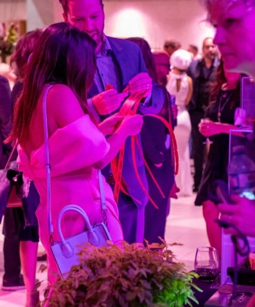 A man and woman are standing at an event, illuminated by pink lighting. The woman is holding several red ribbon-like items, examining them with the man. Various other people are present in the background, and a potted plant and a wine glass are visible in the foreground.