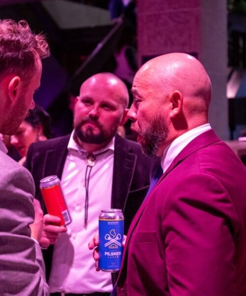 Three men are standing together indoors, engaged in conversation. The man on the left is holding a red can, while the two others are holding blue cans. All three are dressed in suits, with one man wearing a bold pink suit. The background includes a bar and various plants.