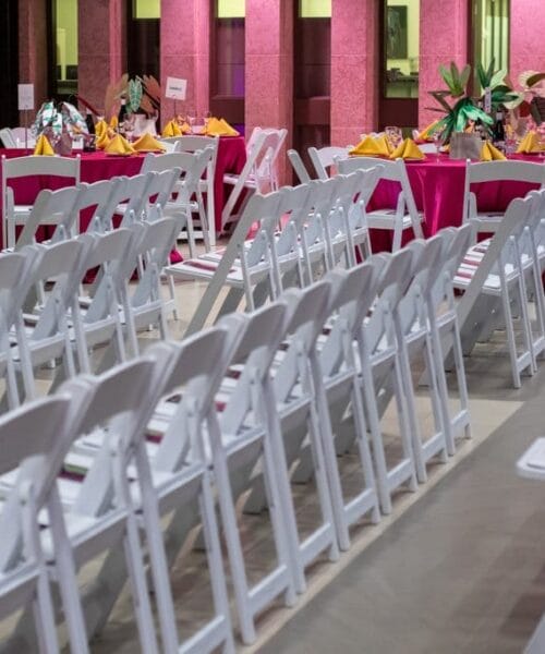Rows of white chairs are arranged in an indoor venue. In the background, round tables covered with bright pink tablecloths are set with yellow napkins and flower centerpieces. A door and walls are partially visible in the distance.