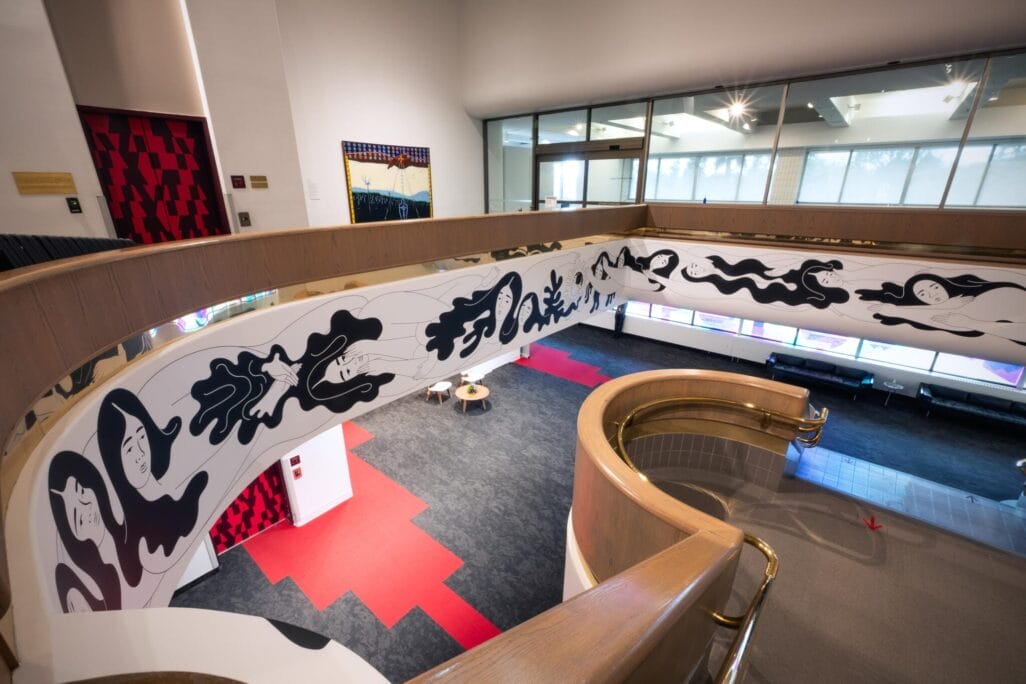 Spiral staircase with a curving banister adorned with bold black and white abstract figures. The staircase leads to an open space with red and gray carpeting, modern art pieces, and a glass-walled meeting room in the background.